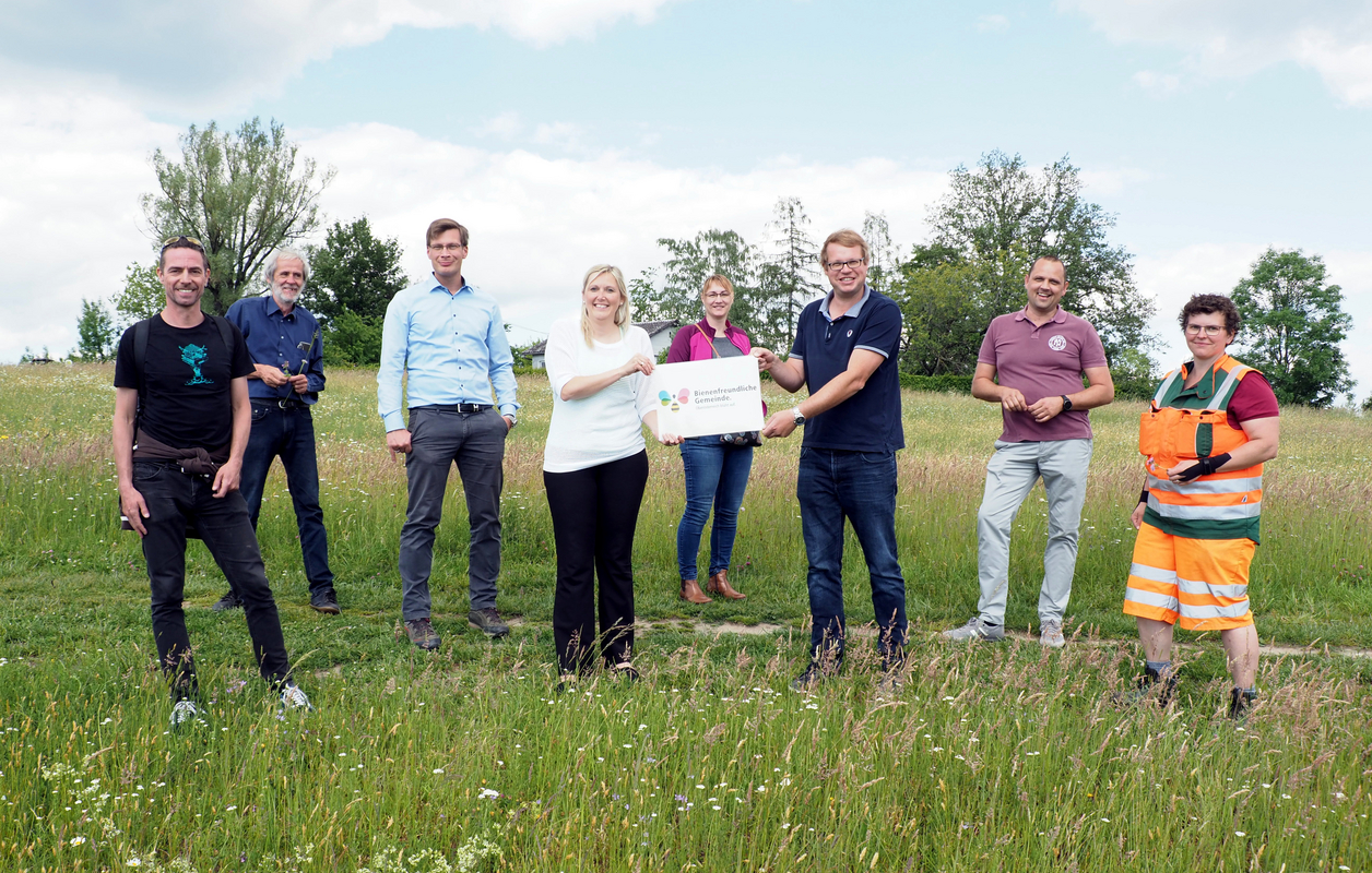 8 Personen in einer Blumenwiese, die vorderen zwei Personen halten ein Schild mit der Aufschrift Bienenfreundliche Gemeinde