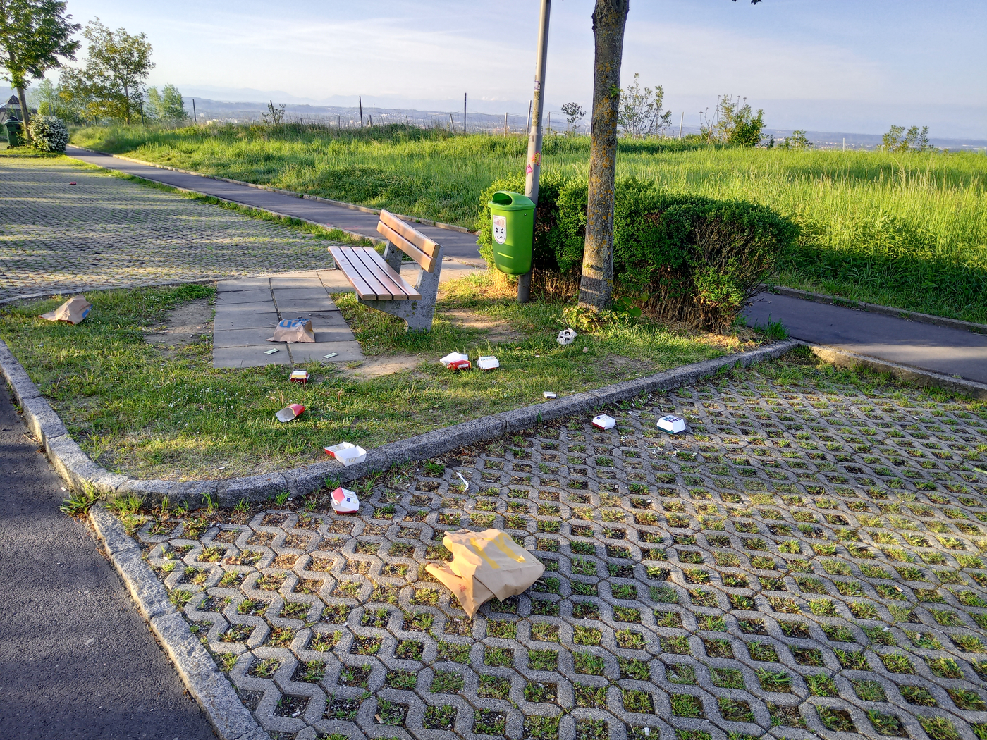 Parkplatz mit einer Bank und einem grünen Mistkübel, Müll liegt am Parkplatz verstreut.